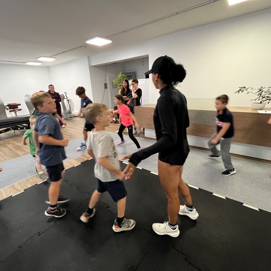 Eine Frau in Trainingskleidung führt eine Gruppe von Kindern durch einen Indoor-Fitnessbereich. Die Kinder rennen auf einem schwarzen Mattenboden herum.