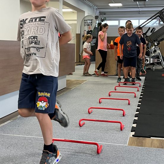 Eine Gruppe von Kindern in einem Fitnessstudio macht eine Hindernisbahn mit roten Stangen, ein Kind springt über die Stangen, während andere zuschauen.