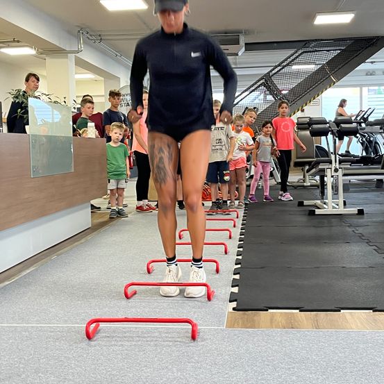 Eine Person in einem Fitnessstudio springt über rote Hürden, während eine Gruppe von Kindern zusieht. Das Fitnessstudio hat verschiedene Trainingsgeräte und eine Treppe.