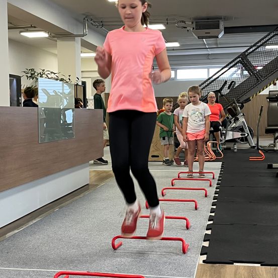 Ein junges Mädchen in einem rosa T-Shirt und schwarzen Leggings springt über rote Hürden in einem Fitnessstudio. Hinter ihr beobachten mehrere Kinder, und eine erwachsene Frau sitzt auf einem Laufband.