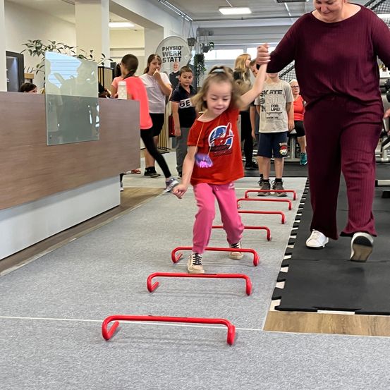 Ein kleines Mädchen in einem roten T-Shirt wird von einer Erwachsenen Frau durch eine Reihe roter Hürden in einem Fitnessstudio geführt. Andere Kinder und Erwachsene sind anwesend.