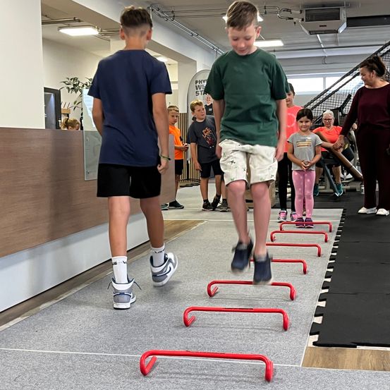 Mehrere Kinder in einem Indoor-Fitnessstudio, eines springt über Hindernisse, andere beobachten. Eine Frau steht in der Nähe, und eine Pflanze ist links.