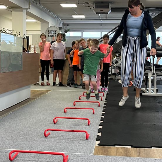 Eine Frau führt einen jungen Jungen mit einem Lächeln über eine Reihe roter Hürden in einem Fitnessstudio. Mehrere Kinder stehen in der Nähe und beobachten.