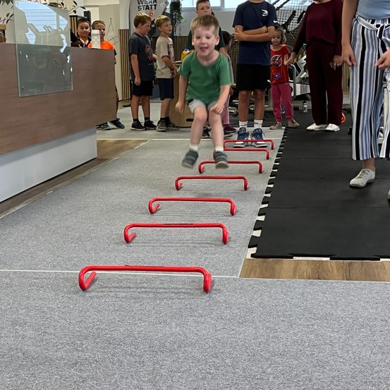 Ein junger Junge springt über rote Hürden in einem Fitnessstudio, während mehrere Kinder und Erwachsene zuschauen. Das Fitnessstudio hat einen grauen Teppich und eine schwarze Matte.