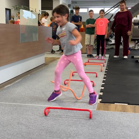 Ein kleines Mädchen in pinken Hosen springt über orange und rote Hürden in einem Fitnessstudio. Andere Kinder und Erwachsene beobachten im Hintergrund.