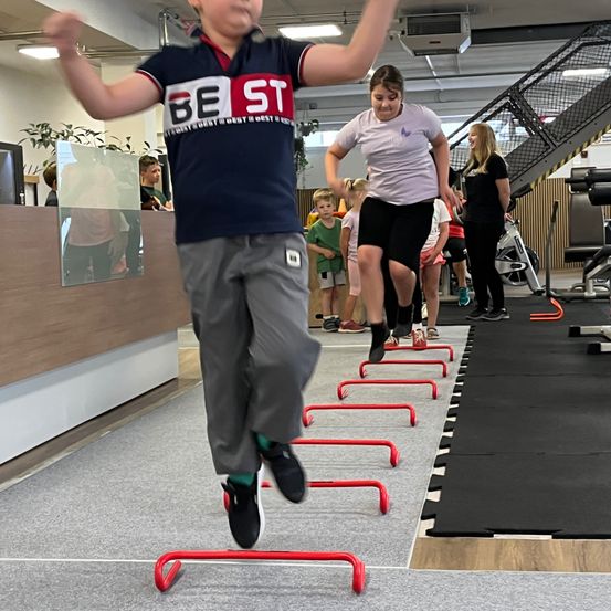 Ein Junge springt über rote Hindernisse in einem Fitnessstudio, er trägt ein blaues Shirt mit weißen Buchstaben. Hinter ihm springt ein Mädchen ebenfalls über die Hindernisse. Andere schauen zu und einige sind auf den Treppen.