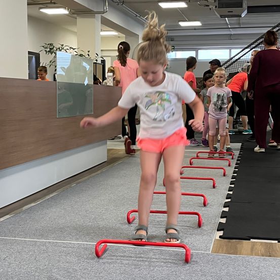 Ein kleines Mädchen springt über rote Hürden auf einem grauen Boden in einem Fitnessstudio. Andere Kinder und Erwachsene sind anwesend und beobachten und nehmen an der Aktivität teil.