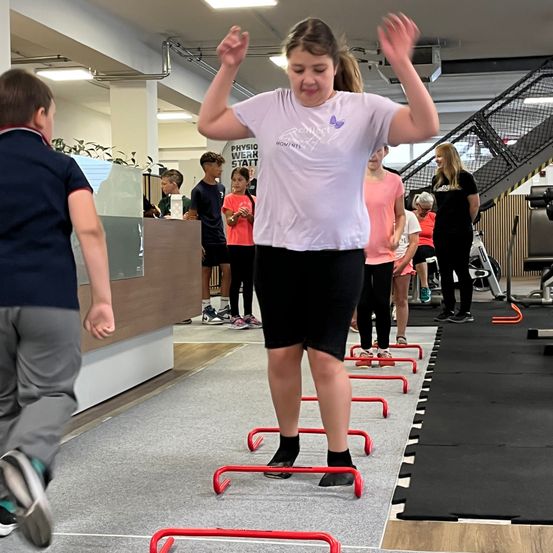 In einem Fitnessstudio springt ein Mädchen über rote Hürden, während andere zuschauen. Hinter ihr stehen eine Gruppe von Kindern und Erwachsenen. Im Hintergrund befinden sich Pflanzen, ein Tresen und eine Treppe.