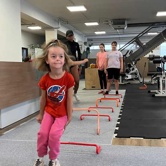 Ein junges Mädchen in einem roten T-Shirt trainiert Geschicklichkeitsübungen mit orangefarbenen und roten Hürden in einem Fitnessstudio, während eine Frau und zwei Mädchen zusehen.