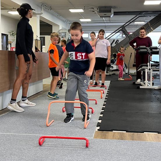 Eine Gruppe von Kindern und Erwachsenen trainiert in einem Fitnessstudio. Ein Junge springt über orange Hürden, während andere herumstehen.