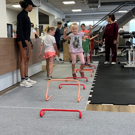 Eine Gruppe von Kindern trainiert im Fitnessstudio eine Hindernisparcours. Eine Frau steht in der Nähe und beobachtet. Sie springen über orange und rote Hürden.