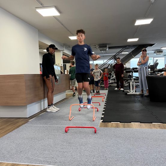 Ein junger Junge springt über Hürden in einem Fitnessstudio. Zuschauer beobachten von der Seite. Das Fitnessstudio hat Geräte und eine Treppe.
