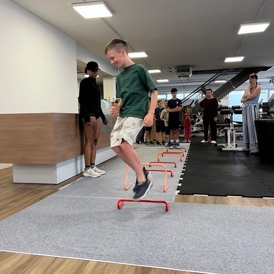Ein Junge in einem grünen T-Shirt springt über rote Hürden in einem Fitnessstudio. Hinter ihm stehen mehrere Menschen und schauen zu, darunter eine Frau in einem schwarzen Shirt und eine Frau in gestreiften Hosen.