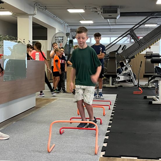 Ein Junge springt über orange Hürden in einem Fitnessstudio, während andere Kinder zuschauen. Das Fitnessstudio verfügt über verschiedene Trainingsgeräte und ein Schild mit der Aufschrift Physio Werk.