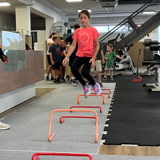 Ein junges Mädchen springt über Hürden in einem Fitnessstudio. Andere Kinder beobachten sie. Das Fitnessstudio hat Trainingsgeräte und ein Schild an der Wand.