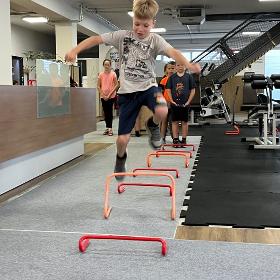 Ein junger Junge springt über Hindernisse in einem Fitnessstudio. Mehrere Kinder beobachten. Das Fitnessstudio hat Trainingsgeräte.