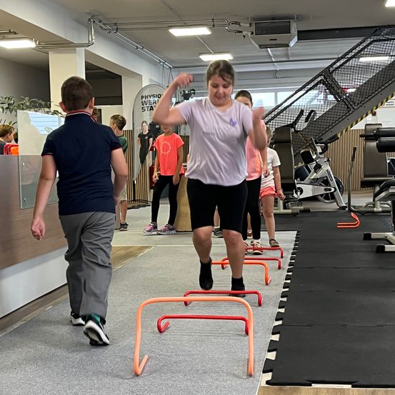 Eine Gruppe von Kindern trainiert in einem Fitnessstudio. Ein Kind springt über orange Hürden, während andere hinter ihr gehen. Das Fitnessstudio verfügt über verschiedene Geräte und eine Treppe.