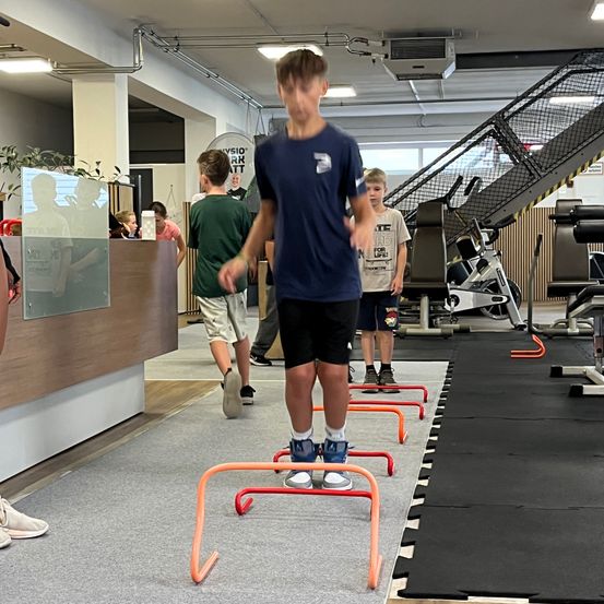 In einem Fitnessstudio steht ein Junge auf orangefarbenen Hürden, während andere in der Nähe gehen. Das Fitnessstudio hat einen schwarzen Boden, Trainingsgeräte und eine Treppe.