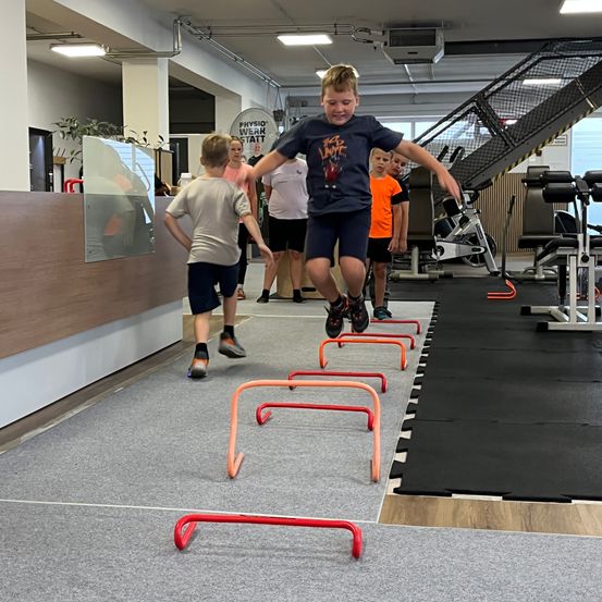 Eine Gruppe von Kindern springt in einem Fitnessstudio über Hürden. Ein Junge führt und springt voller Begeisterung. Andere folgen hinter ihm und warten auf ihren Zug. Fitnessgeräte und eine Treppe sind im Hintergrund zu sehen.