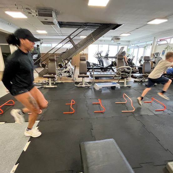 Zwei Personen führen in einem Fitnessstudio Agilitätsübungen durch. Eine springt über orangefarbene Hütchen, während die andere einen ähnlichen Weg verfolgt. Das Fitnessstudio ist mit verschiedenen Trainingsgeräten und einer Treppe ausgestattet.