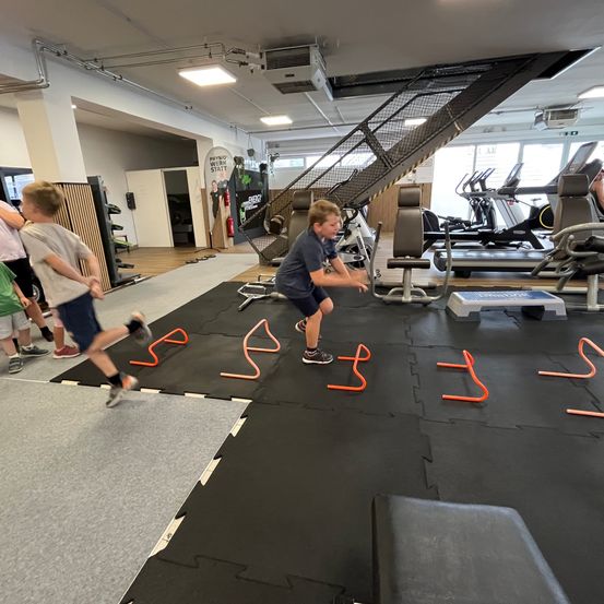 In einem Fitnessstudio laufen zwei Jungen auf einer Matte mit orangefarbenen Hütchen. Sie sind von Fitnessgeräten und einer Treppe umgeben.