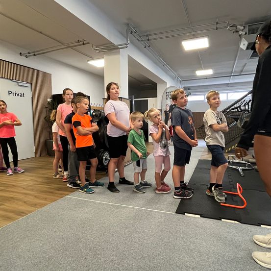 Eine Gruppe von Kindern und Erwachsenen steht in einem Fitnessstudio. Eine Frau in Schwarz führt sie an. Sie tragen verschiedene farbige T-Shirts und Sneakers. Ein schwarzer Teppich liegt auf dem Boden.
