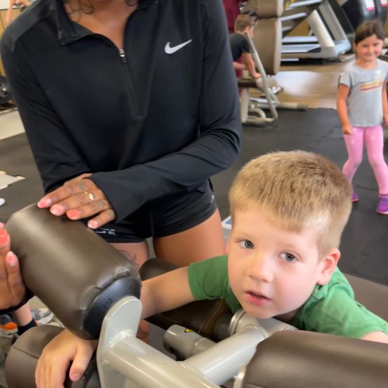 Eine Frau in einem schwarzen Nike-Langarmshirt hilft einem kleinen Jungen, eine Hantelbank in einem Fitnessstudio zu benutzen. Ein kleines Mädchen steht in der Nähe.