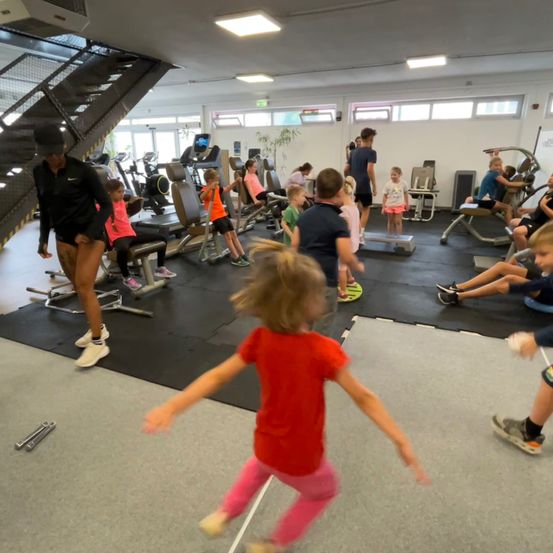 Ein Fitnessstudio mit verschiedenen Trainingsgeräten. Eine Frau in schwarzer Kleidung steht. Kinder laufen und spielen im Vordergrund.