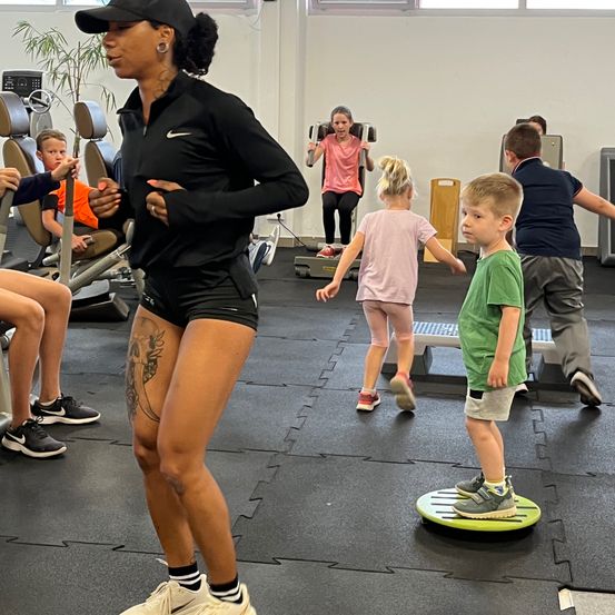 Eine Frau in Trainingskleidung leitet eine Gruppe von Kindern in einem Fitnessstudio, wobei sie Fitness und Spaß in den Vordergrund stellt. Die Kinder nehmen an verschiedenen Übungen teil, einige auf Balancebrettern.