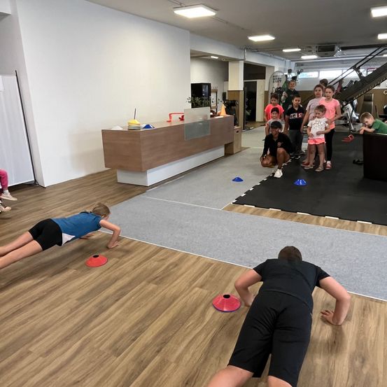 Eine Gruppe von Kindern und Erwachsenen macht Liegestütze auf einem Holzboden in einem Fitnessstudio, einige auf einer Matte und andere beobachten. Bunte Kegel sind auf dem Boden platziert.
