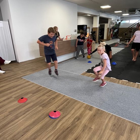 Kinder spielen ein Spiel in einem Fitnessstudio, rennen und springen über Kegel. Ein Mädchen hebt einen Ball auf.