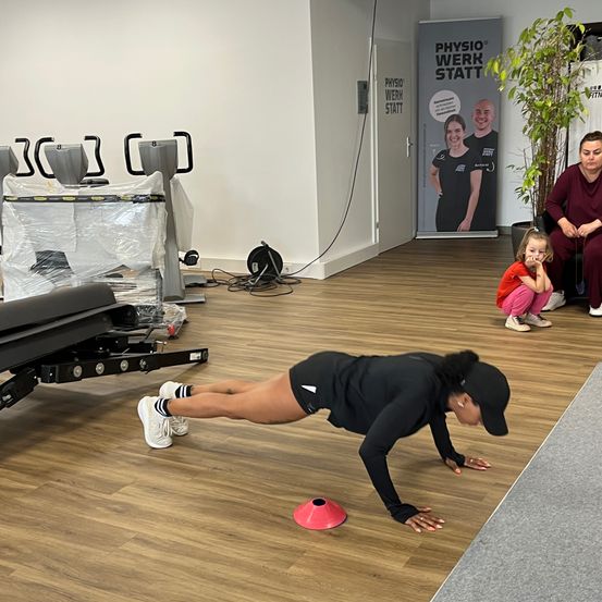 Eine Frau führt Liegestütze in einem Fitnessstudio durch, vor ihr steht ein rosafarbener Kegel. In der Nähe sitzen eine Frau und ein Kind und beobachten. Fitnessgeräte und ein Banner für Physiotherapie sind im Hintergrund zu sehen.