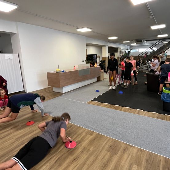 In einem Fitnessstudio beschäftigen sich mehrere Personen mit Trainingsaktivitäten. Zwei Personen liegen auf dem Boden mit roten Kegeln vor ihnen. Andere stehen herum, möglicherweise beobachten sie oder warten auf ihren Zug. Ein Tresen ist im Hintergrund zu sehen.