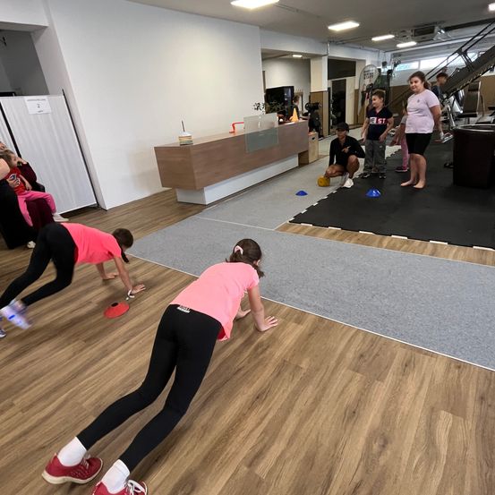 Ein Fitnessstudio voller Kinder, die Sport treiben. Zwei Mädchen machen Liegestütze, während andere zuschauen.