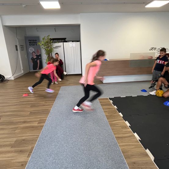 Kinder in einem Fitnessstudio, die auf einem Teppich laufen, eines in einem schwarzen Shirt und das andere in einem rosa Shirt. Eine Erwachsene sitzt im Hintergrund, und es gibt ein Plakat an der Wand.