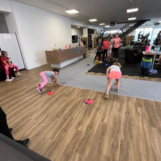Mehrere Kinder in einem Fitnessstudio nehmen an einem Spiel mit roten Scheiben auf dem Boden teil. Zwei Kinder beugen sich vor, möglicherweise um eine Scheibe aufzuheben.