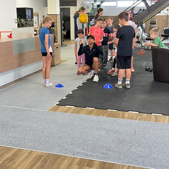 In einem Fitnessstudio unterrichtet ein Mann eine Gruppe von Kindern. Sie stehen auf einer schwarzen Matte mit blauen Hütchen. Die Kinder stehen in einer Reihe und scheinen dem Mann zuzuhören.