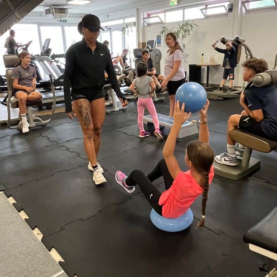Ein Fitnesskurs mit Kindern und Erwachsenen, ein Kind hält einen blauen Ball, eine Frau gibt Anweisungen, während ein anderes Kind zuschaut.