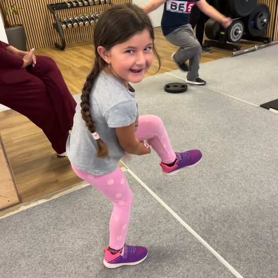 Ein kleines Mädchen im Fitnessstudio, lächelnd, mit zwei Personen im Hintergrund, eine hebt Gewichte und die andere sitzt auf dem Boden.