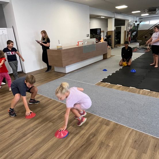 Eine Gruppe von Kindern und Erwachsenen spielt mit farbigen Scheiben auf einem Holzboden in einem Fitnessstudio. Eine Frau steht in der Nähe des Tresens und ein Mädchen macht Yoga auf dem Boden.