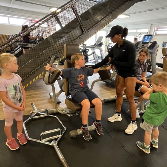 In einem Fitnessstudio hilft eine Frau einem Jungen auf einem Trainingsstuhl. Drei Kinder stehen um ihn herum und schauen zu. Im Hintergrund ist eine Treppe mit einer Person auf einem Laufband zu sehen.
