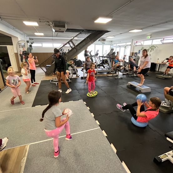In einem Fitnessstudio nimmt eine Gruppe von Kindern unter Aufsicht von Erwachsenen an einem Trainingskurs teil. Ein Kind steht auf einem Balancebrett, während andere Trainingsbälle halten. Das Fitnessstudio ist gut beleuchtet und mit verschiedenen Trainingsgeräten ausgestattet.