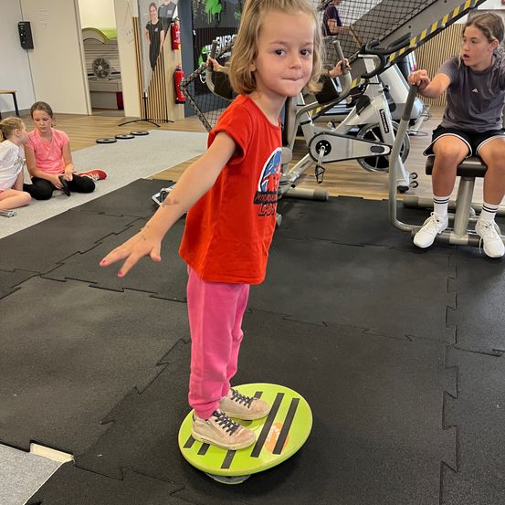 Ein kleines Mädchen balanciert auf einem Wobble-Board im Fitnessstudio. Andere Kinder sitzen und beobachten.