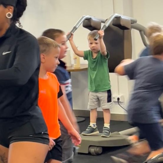 Eine Frau in Schwarz führt eine Gruppe von Kindern in einem Fitnessstudio. Ein Junge in Grün hält sich an einem Laufband fest, während andere zuschauen.