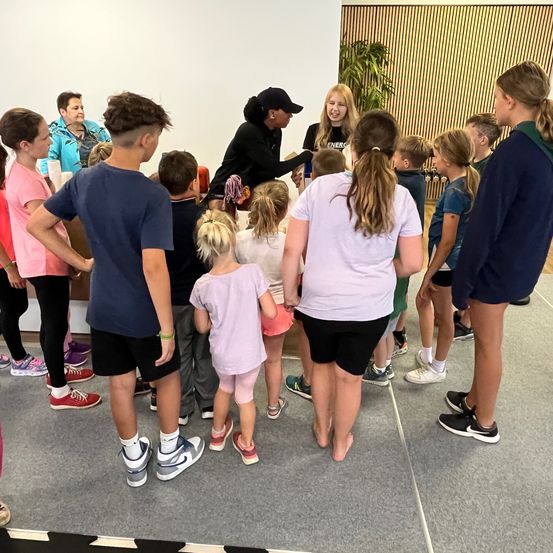 Eine Gruppe von Kindern und Erwachsenen steht in einem Kreis, wahrscheinlich in einem Klassenzimmer. Einige tragen Turnschuhe, und eine Frau in einer Mütze gibt etwas aus.