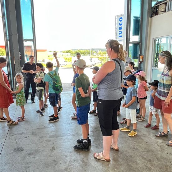 Eine Gruppe von Erwachsenen und Kindern steht in einem Gebäude, wahrscheinlich einer Feuerwache. Sie alle schauen nach vorne, wahrscheinlich jemandem zuhörend. Das Gebäude hat eine Glastür und Fenster.