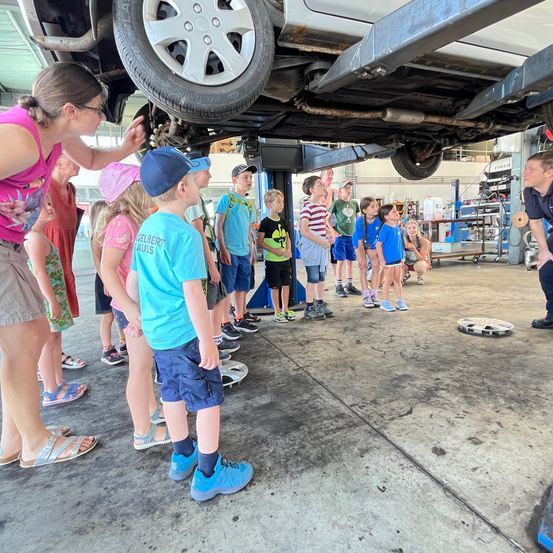 In einer Werkstatt stehen Kinder um ein auf Bockwerken gehobenes Auto. Ein Erwachsener zeigt auf das Fahrzeug und erklärt den Kindern etwas.