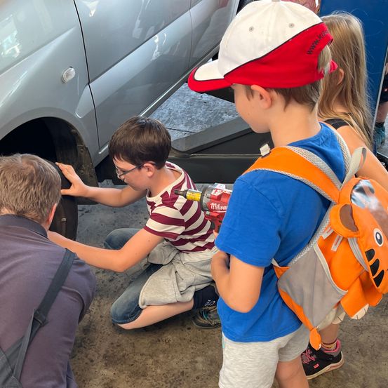 Eine Gruppe von Kindern versammelt sich um ein Fahrzeug, ein Kind benutzt ein Werkzeug, um an der Reifen zu arbeiten. Der Junge im Vordergrund trägt ein gestreiftes Shirt und eine Brille. Der Junge neben ihm trägt ein blaues Shirt und einen orangefarbenen Rucksack.