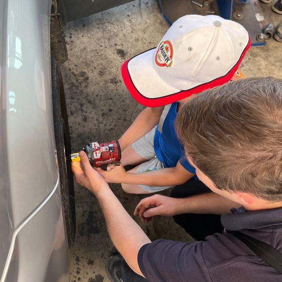 Zwei Personen arbeiten an einem Auto, eine benutzt ein Werkzeug am Reifen, während die andere eine Bohrmaschine hält. Einer trägt ein blaues Hemd und eine Mütze mit Logo.