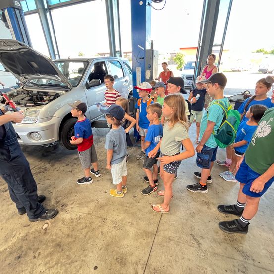 Eine Gruppe von Kindern in einer Garage, die einen Erwachsenen dabei beobachten, wie er den Motor eines Autos untersucht. Sie stehen nah am Auto, dessen Motorhaube geöffnet ist.
